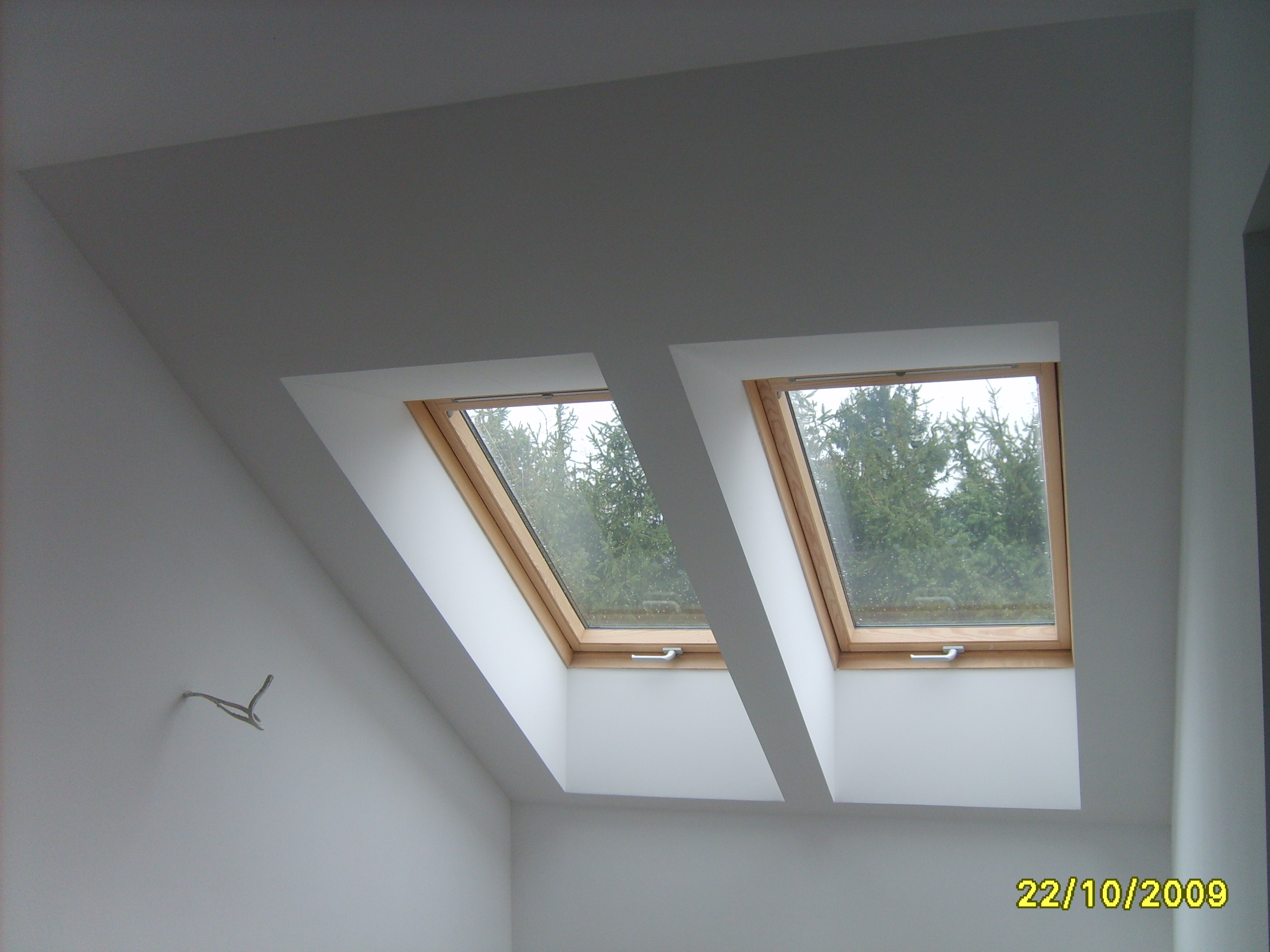 Poddasze z dwoma oknami dachowymi w drewnianych ramach, świeżo po malowaniu na biało, widoczny wystający kabel ze ściany.